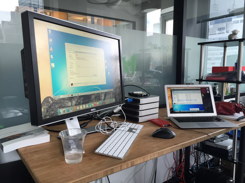 A Dell monitor, MacBook laptop, keyboard, mouse, and stack of external drives on a desk, with a glass of water and a book about fuzzing