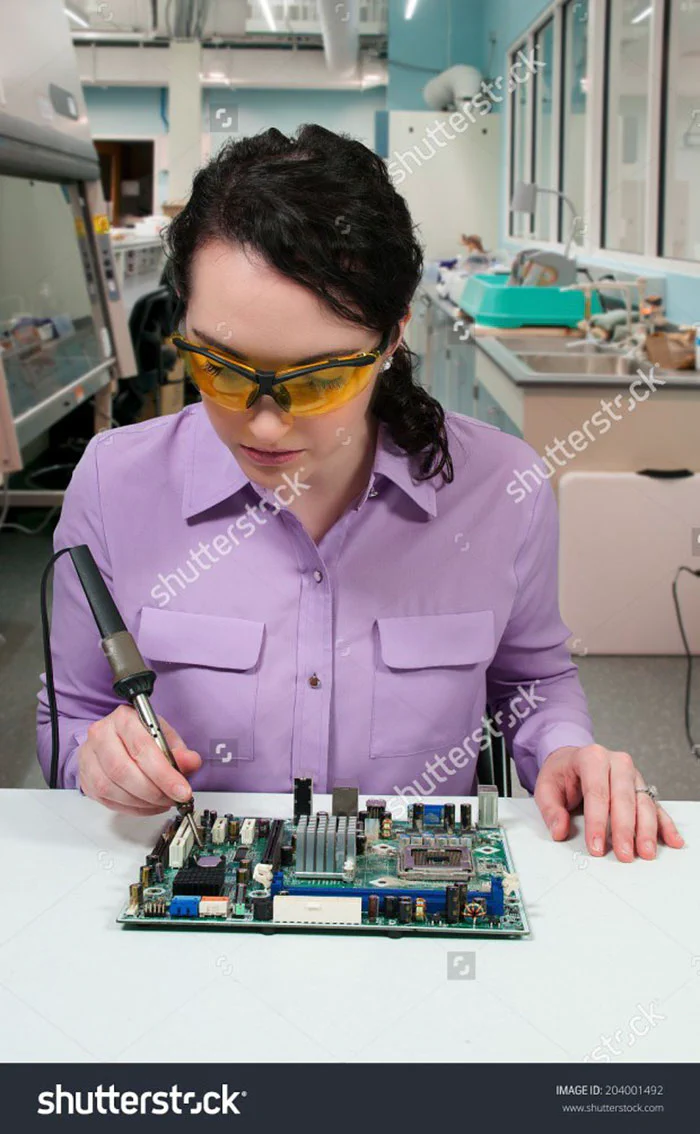 Stock photo of a woman wearing safety goggles while soldering a circuit board with no other protective gear